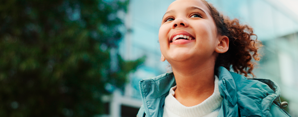 Menina sorrindo alegremente para o alto, com casaco azul, simbolizando esperança e futuro
