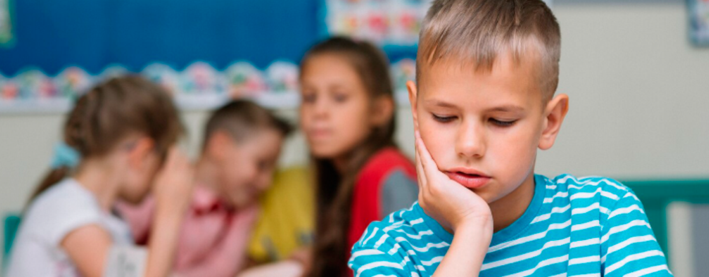 Menino com camiseta azul e branca, visivelmente pensativo e isolado em uma sala de aula, enquanto outras crianças interagem ao fundo. Pode indicar bullying ou problemas de socialização.